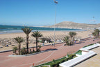 Agadir Beach Club Hotel beachfront view showing sandy beach with sun umbrellas, palm trees, and a paved promenade under clear blue sky