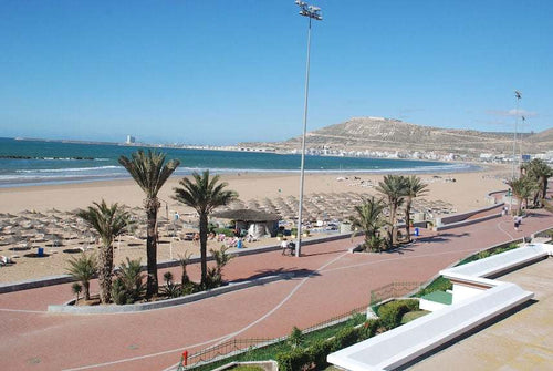 Agadir Beach Club Hotel beachfront view showing sandy beach with sun umbrellas, palm trees, and a paved promenade under clear blue sky