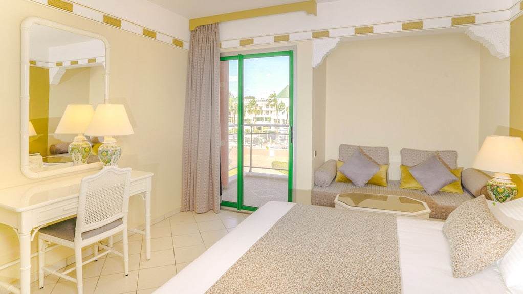 Agadir Beach Club Hotel room with cream walls, white tiled floor, beige bedspread, decorative lamps, and green-framed balcony door