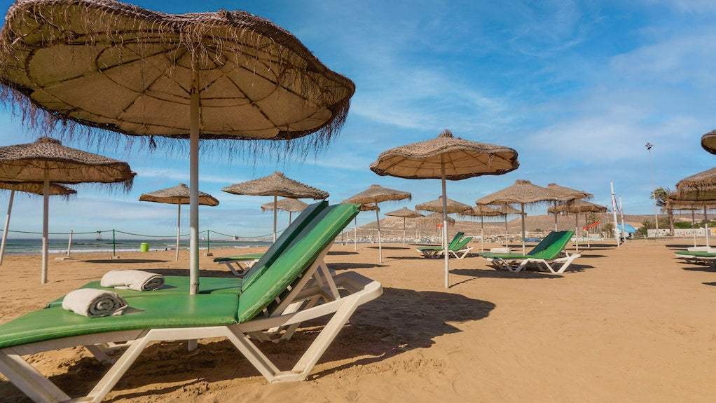Agadir Beach Club Hotel sandy beach with green lounge chairs and thatched umbrellas under a blue sky