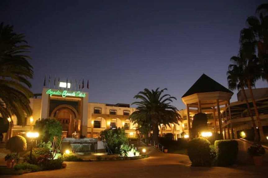 Agadir Beach Club Hotel illuminated at dusk with palm trees, garden landscaping, and a fountain in front