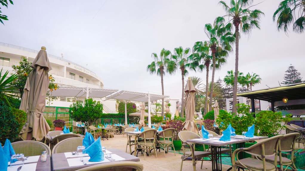 Outdoor dining area at Agadir Beach Club Hotel with beige wicker chairs, tables set with blue napkins, and palm trees