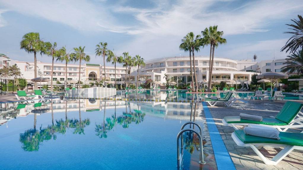 Agadir Beach Club Hotel with large outdoor pool, green lounge chairs, palm trees, and white building under clear sky