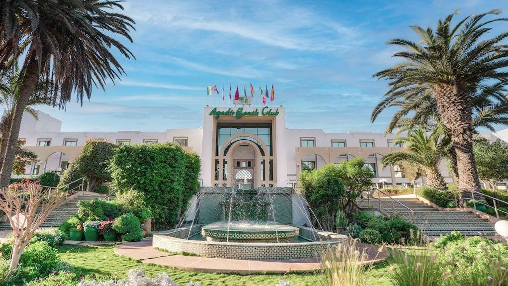 Agadir Beach Club Hotel with white facade, Moroccan-style arch entrance, surrounded by palm trees and green landscaping