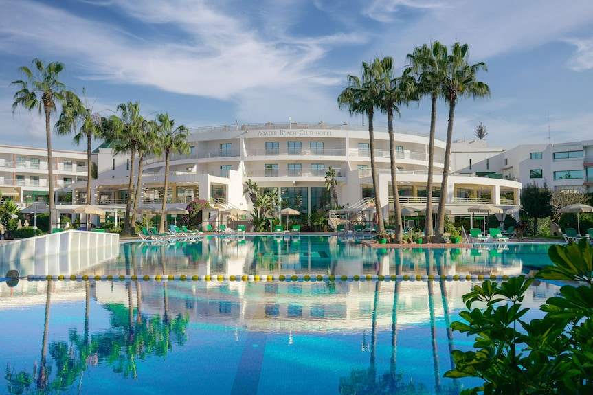 Agadir Beach Club Hotel large white resort building with palm trees and a blue swimming pool reflecting the sky and hotel