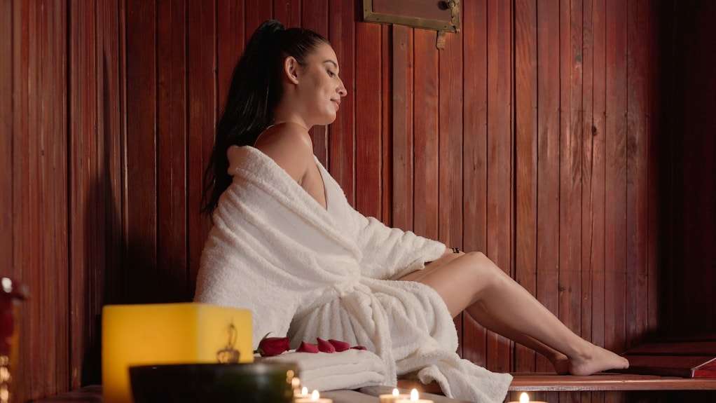 Woman in white bathrobe sitting inside a wooden sauna room with lit candles and rose petals, relaxing at Agadir Beach Club Hotel