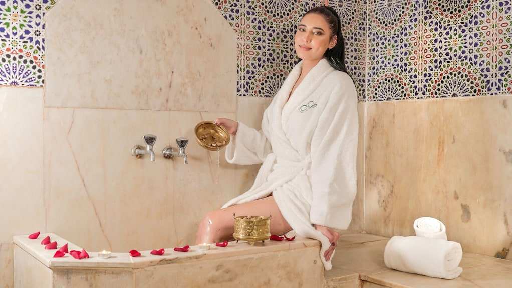Woman in white bathrobe sitting on marble edge in a traditional Moroccan hammam with colorful patterned tiled walls relaxing