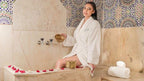 Woman in white bathrobe sitting on marble edge in a traditional Moroccan hammam with colorful patterned tiled walls relaxing