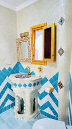 Bathroom sink area with geometric blue and white tilework, wooden framed mirror and window, reflecting Alhambra Ryad & Spa style