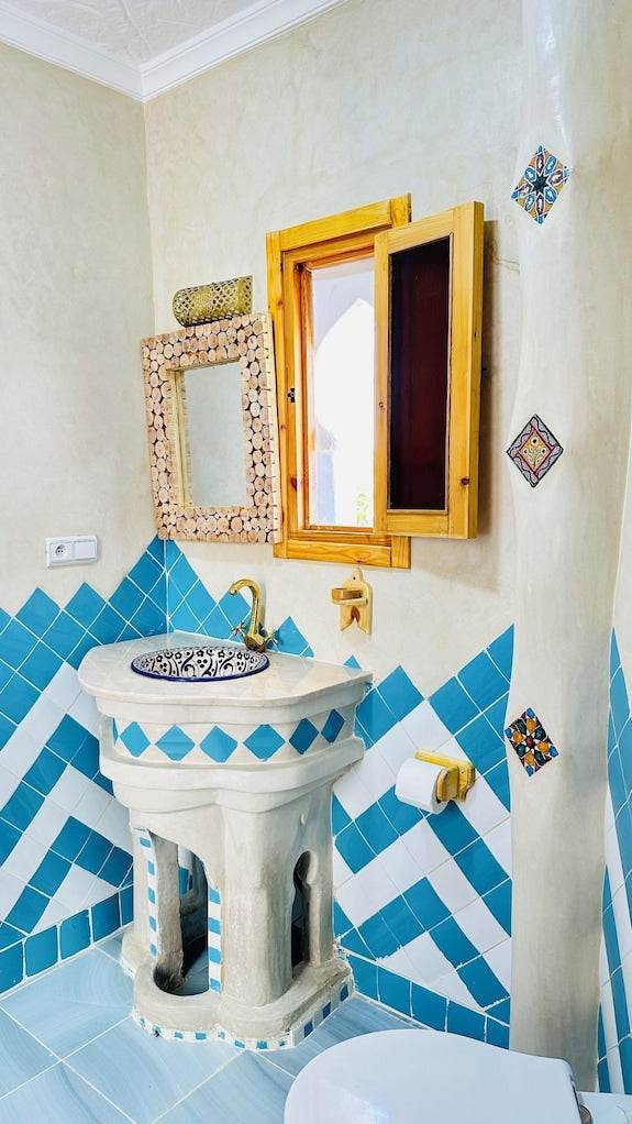 Bathroom sink area with geometric blue and white tilework, wooden framed mirror and window, reflecting Alhambra Ryad & Spa style