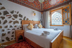 Alhambra Ryad & Spa bedroom with carved wooden bed, white linens, patterned pillows, stone and white wall, ornate ceiling and arched window