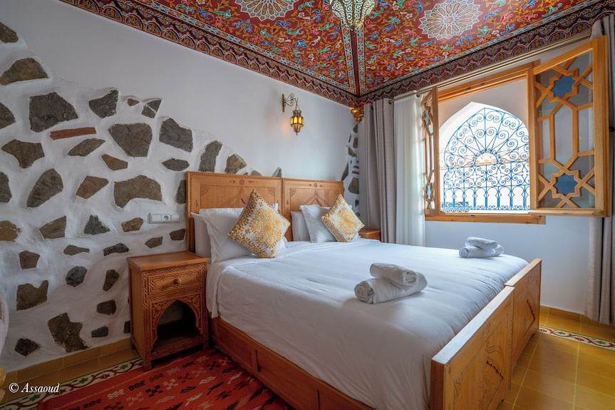 Alhambra Ryad & Spa bedroom with carved wooden bed, white linens, patterned pillows, stone and white wall, ornate ceiling and arched window