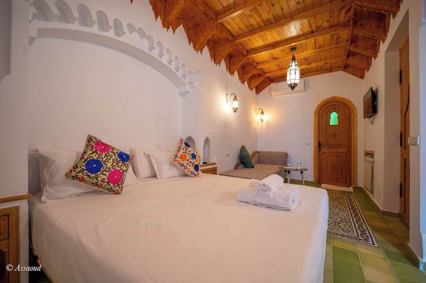 Alhambra Ryad & Spa bedroom with wooden ceiling, white walls, colorful floral cushions, green tiled floor, and wooden door