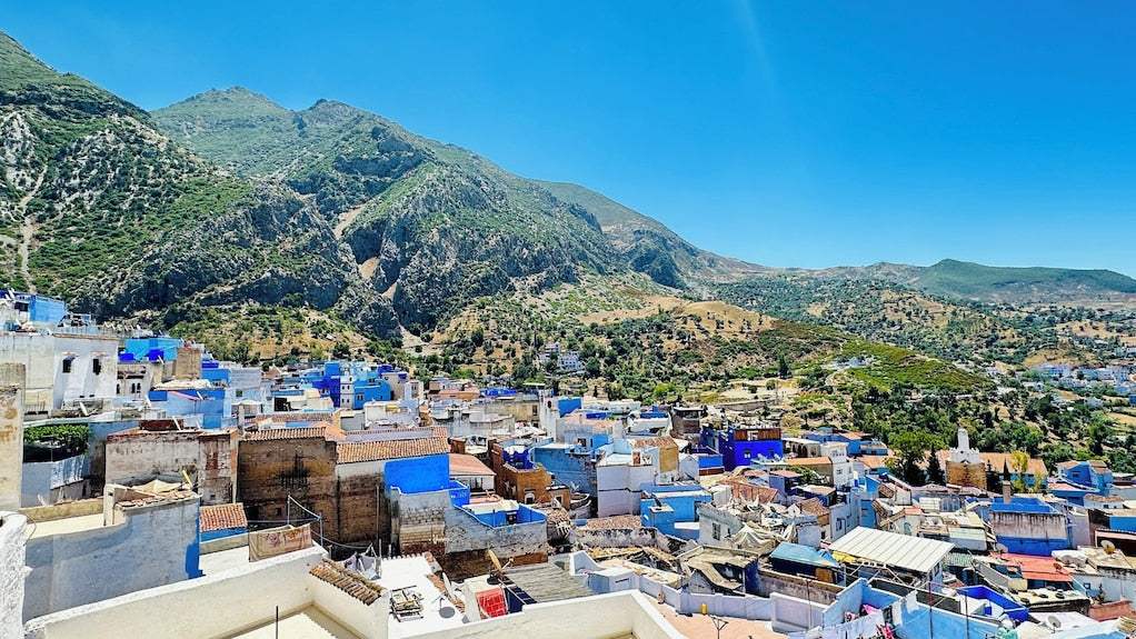 Panoramic view of Chefchaouen with blue and white painted buildings nestled among green mountains under a clear blue sky at Alhambra Ryad & Spa