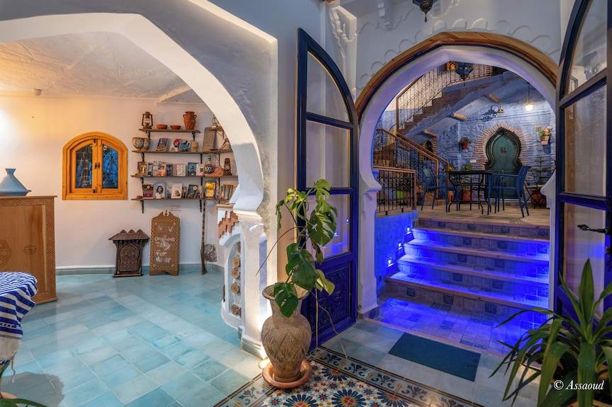Alhambra Ryad & Spa interior with blue tiled floors, white walls, arched doorways, wooden shelves, and vibrant blue-lit staircase leading to an outdoor seating area