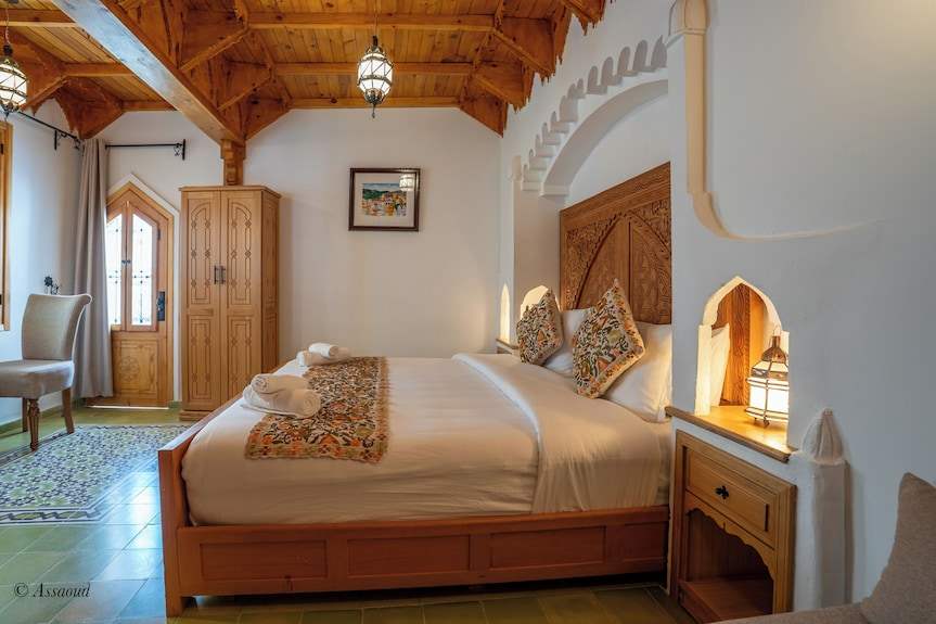 Alhambra Ryad & Spa room with wooden bed frame and headboard, white linens, patterned cushions and runner, wooden ceiling, and Moroccan tile floor