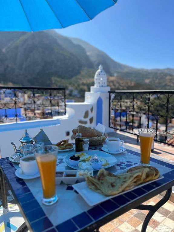 Alhambra Ryad & Spa rooftop breakfast setup with traditional Moroccan tea, orange juice, bread, and scenic mountain views in Chefchaouen