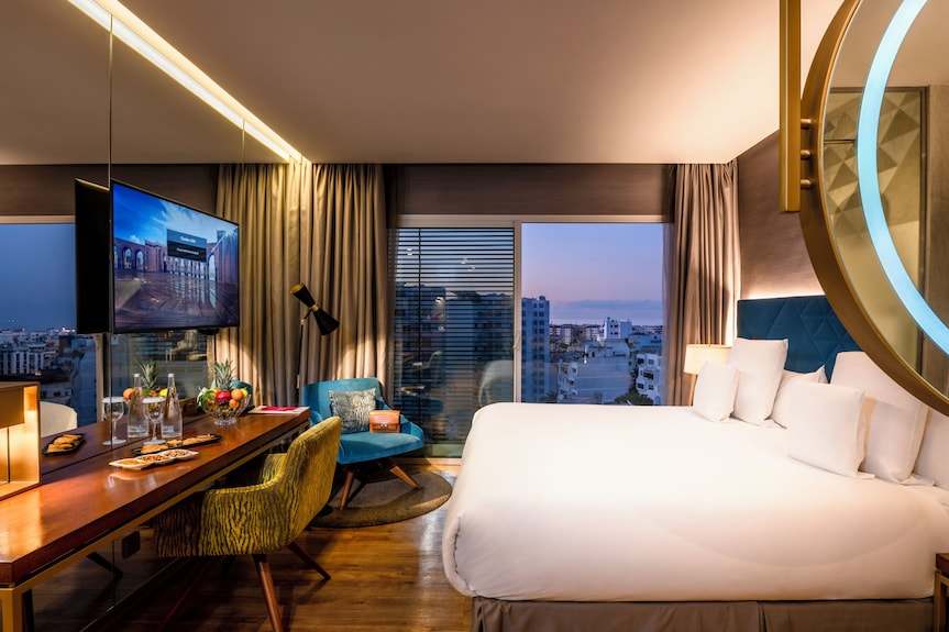Barcelo Anfa Casablanca hotel room with white king bed, wooden furnishings, blue chairs, and city views