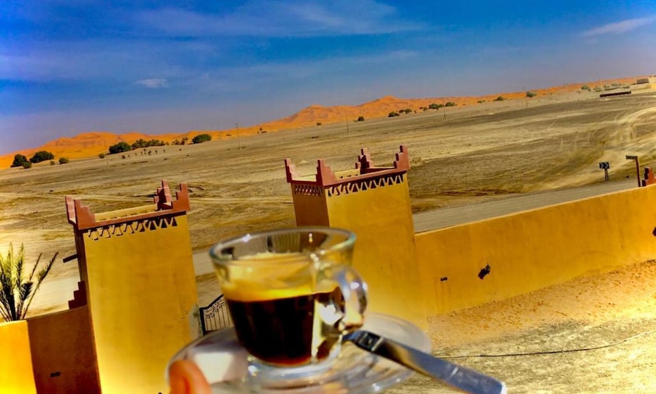 Berber Palace Hotel view featuring a clear blue sky, sandy landscape, and traditional yellow desert walls with a glass cup of black coffee in the foreground