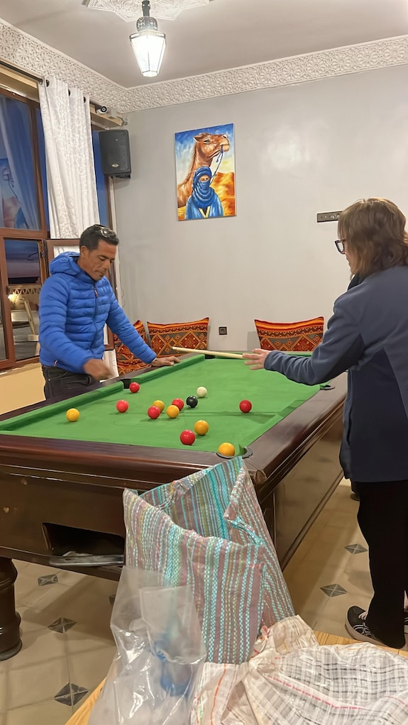 Berber Palace Hotel indoor pool table with green felt top, two people playing billiards, colorful cushions, and camel painting on wall
