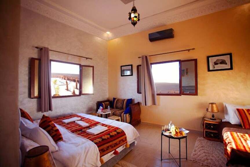 Hotel bedroom at Berber Palace Hotel with beige and orange walls, twin beds featuring patterned red and orange blankets, and a small round table with a fruit platter.