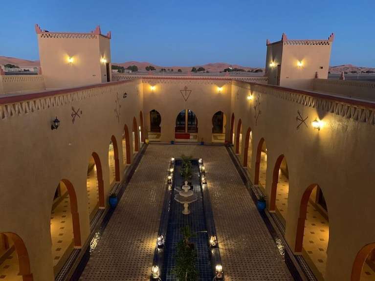 Berber Palace Hotel courtyard at dusk with traditional Moroccan architecture, warm beige walls, arched doorways, and illuminated lanterns
