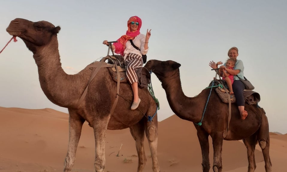 Two brown camels with saddles carrying smiling women and a child in desert sand dunes during sunset at Berber Palace Hotel