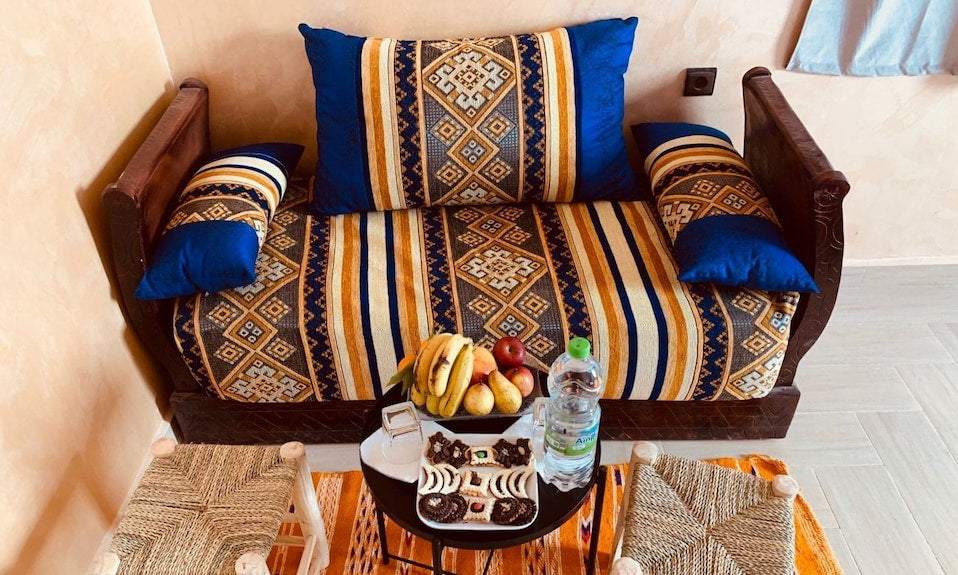 Berber Palace Hotel wooden sofa with blue and beige patterned cushions, accompanied by two woven stools and a small table with fruit and water bottle