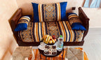 Berber Palace Hotel wooden sofa with blue and beige patterned cushions, accompanied by two woven stools and a small table with fruit and water bottle