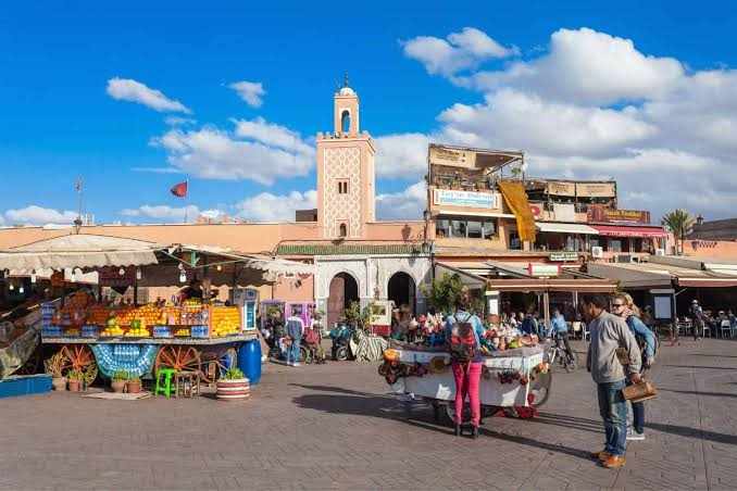 Street market in Marrakech with colorful fruit stalls, traditional Moroccan architecture, and people shopping under a bright blue sky featuring Booking Flight Tickets