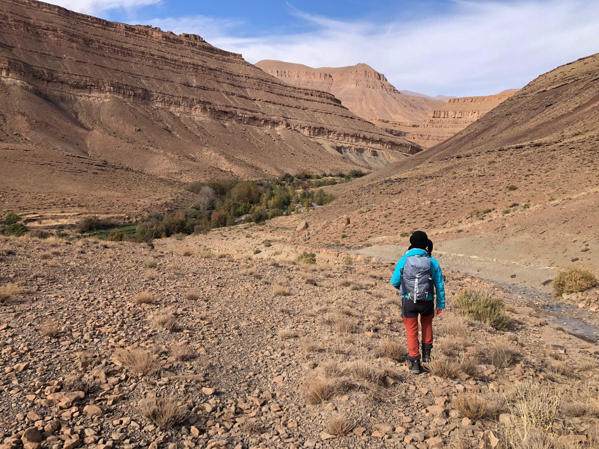 Boumalne Dades Tour featuring a person hiking through a rocky, arid valley with layered brown mountain cliffs and sparse vegetation