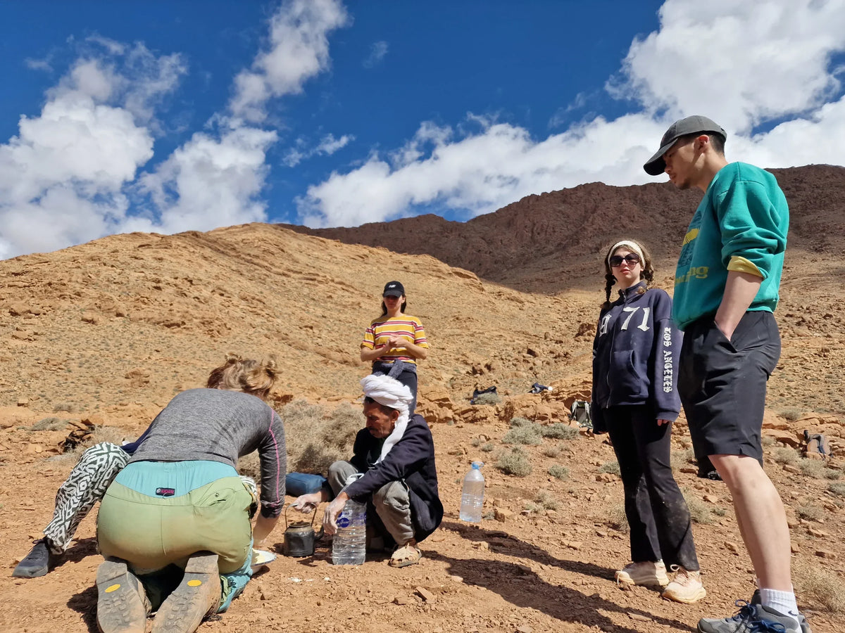 Boumalne Dades Tour group with diverse hikers in casual outdoor clothing gathered around traditional Berber man in rocky mountain landscape