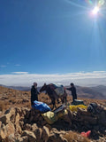 Boumalne Dades Tour showing two people with a loaded mule on a rocky mountain landscape under a bright sun, highlighting outdoor adventure and traditional transport