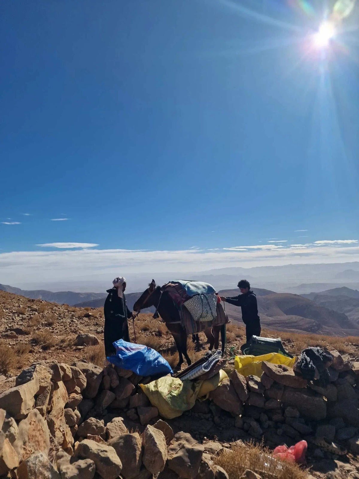 Boumalne Dades Tour showing two people with a loaded mule on a rocky mountain landscape under a bright sun, highlighting outdoor adventure and traditional transport