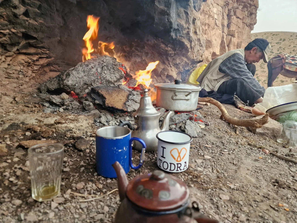 Outdoor Moroccan tea setup with metal teapot, blue and white mugs near open fire and man preparing tea on Boumalne Dades Tour