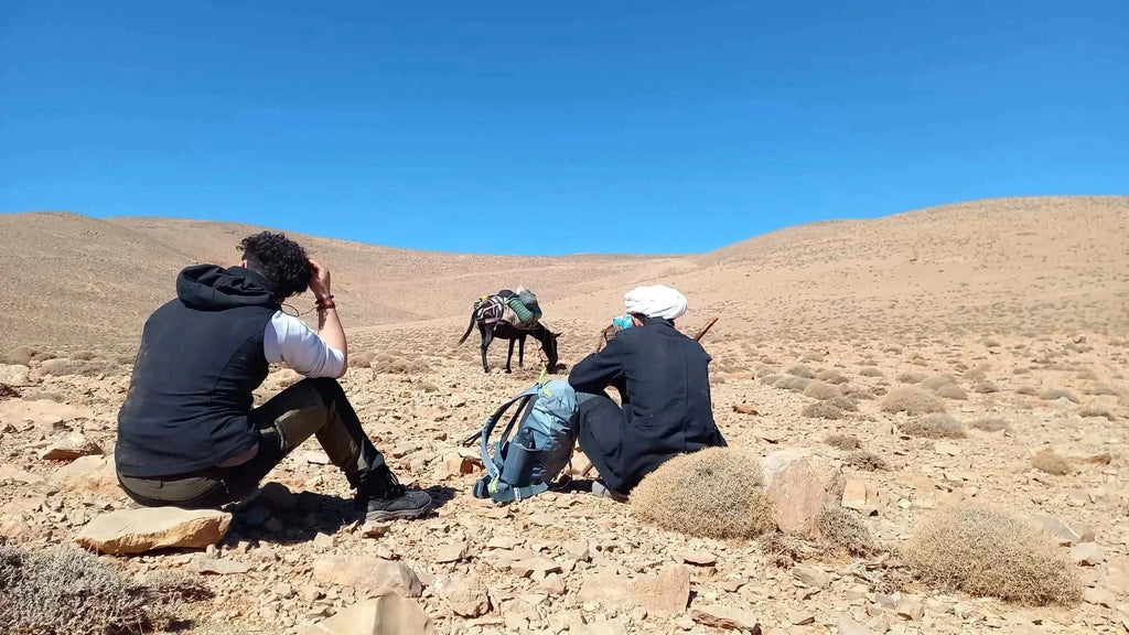 Boumalne Dades Tour showing two hikers resting on rocky terrain with a donkey carrying supplies under a clear blue sky