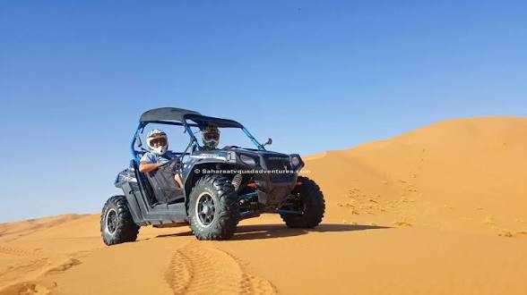 Off-road black and grey dune buggy with two riders wearing helmets on golden sand dunes under clear blue sky for Buggy Rental in Merzouga.