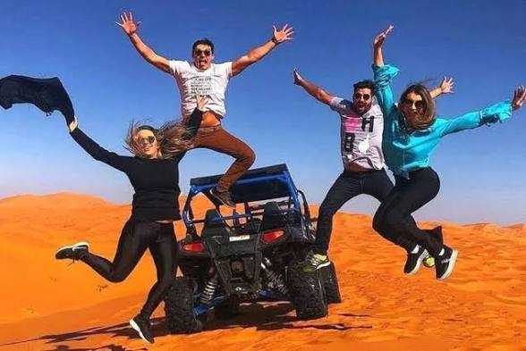 Group of four people jumping with joy next to a blue off-road vehicle on red sand dunes under clear sky, capturing the Merzouga Sahara Desert adventure.