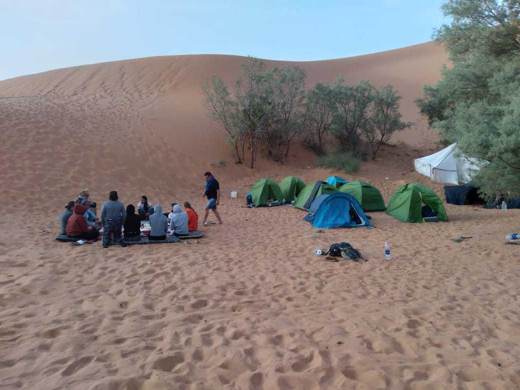 Camel Trekking desert camp scene with group of people sitting on sand near green and blue tents against sand dunes and trees
