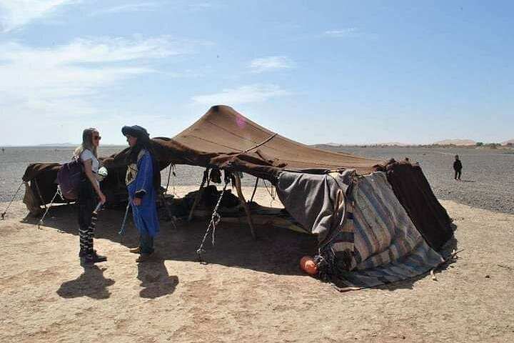Camel trekking scene with a traditional brown desert tent and two people dressed for desert travel standing nearby on arid ground under a blue sky