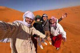 Camel trekking with four people wearing traditional desert clothing and headscarves on orange Sahara desert dunes smiling and waving