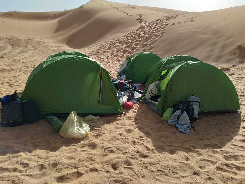 Three green dome tents pitched on sandy desert ground surrounded by camping gear near sandy dunes during a Camel Trekking adventure