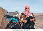 Camel trekking guide wearing a colorful traditional robe and pink headscarf in the desert near camel saddles and equipment