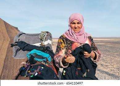Camel trekking guide wearing a colorful traditional robe and pink headscarf in the desert near camel saddles and equipment
