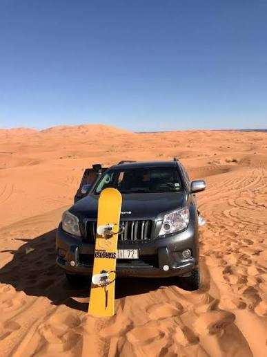SUV in dark grey parked on golden sand dunes with a yellow snowboard standing upright in front, depicting Camel Trekking in the Sahara Desert adventure.