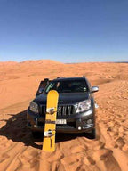 SUV in dark grey parked on golden sand dunes with a yellow snowboard standing upright in front, depicting Camel Trekking in the Sahara Desert adventure.