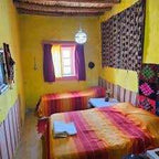 Traditional Moroccan guest room with yellow walls, two beds with red-orange striped bedding, and decorative textiles, ideal for Camel Trekking in the Sahara Desert accommodations.