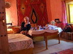Cozy desert guest room with warm orange walls, traditional patterned textiles, wooden furniture, and a person sitting by a table enjoying tea, evoking the spirit of Camel Trekking in the Sahara Desert.
