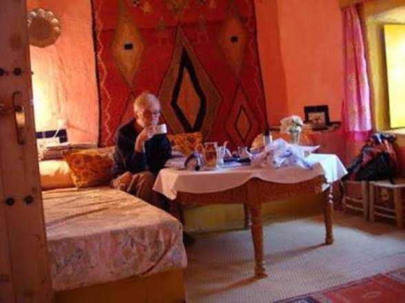 Cozy desert guest room with warm orange walls, traditional patterned textiles, wooden furniture, and a person sitting by a table enjoying tea, evoking the spirit of Camel Trekking in the Sahara Desert.