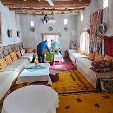 Traditional Moroccan sitting room with white walls, wooden ceiling beams, colorful rugs, cushions, and low seating, perfect for Camel Trekking in the Sahara Desert accommodations.